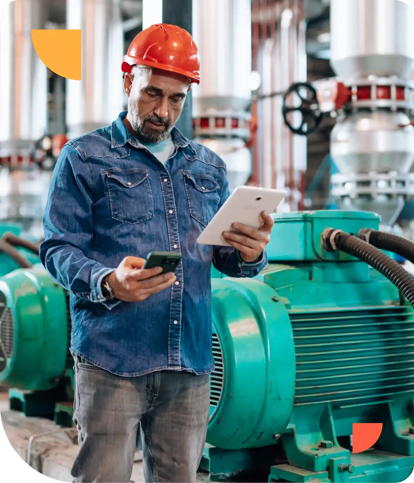 Man in a red hard hat and denim shirt using a tablet and smartphone in an industrial setting with machinery in the background.