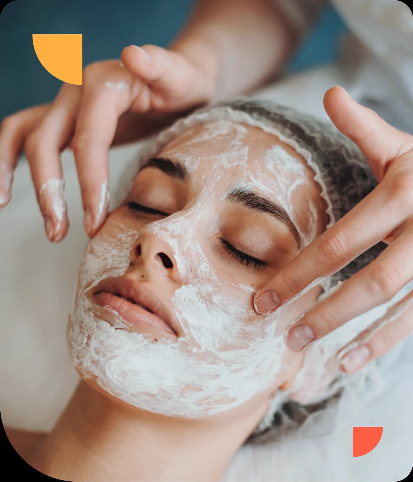 Person receiving a facial treatment, eyes closed, with a creamy mask applied, wearing a hair cap, and hands gently massaging their face.