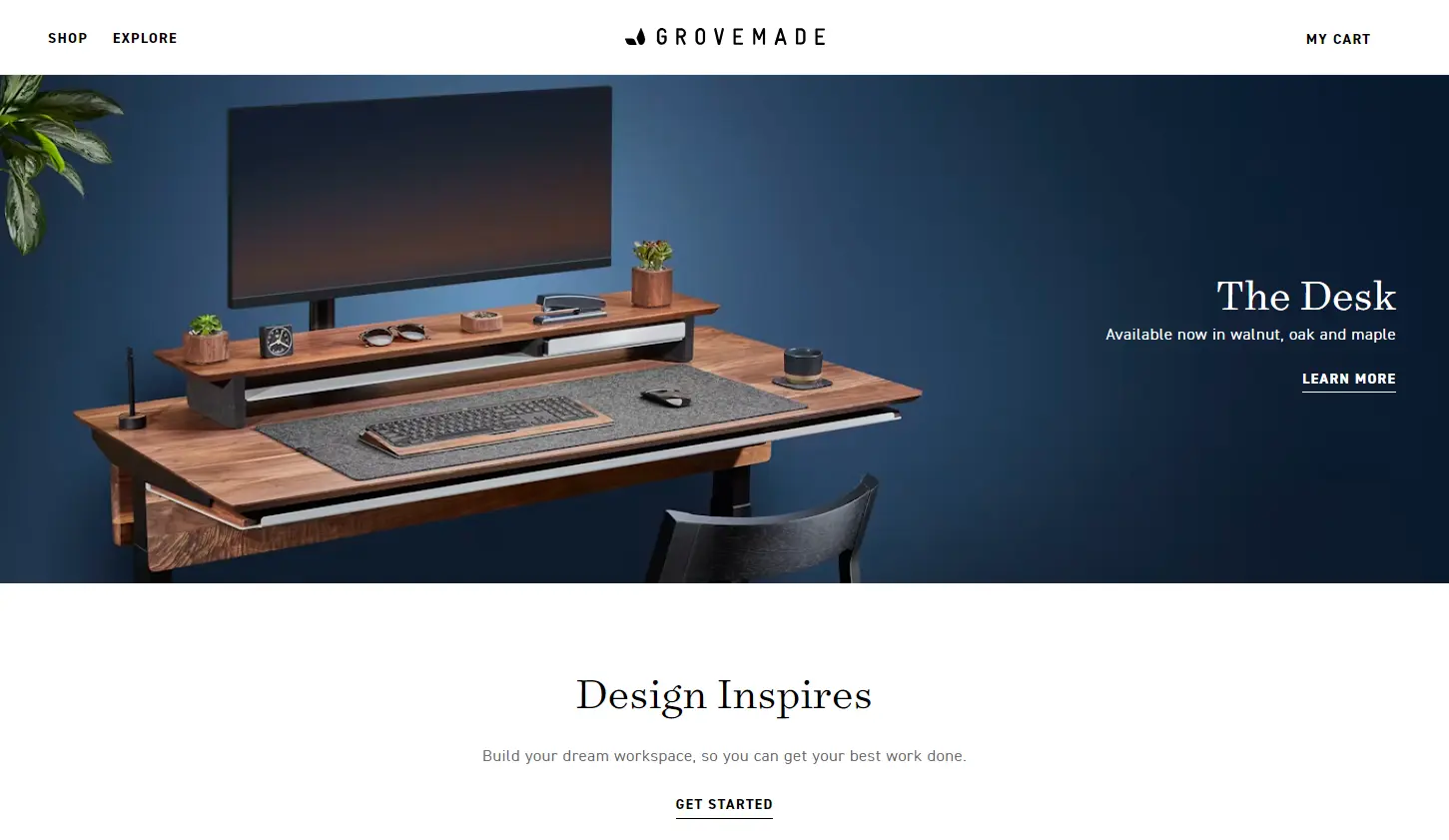 A modern desk setup with a monitor, keyboard, mouse, and plants on a wooden surface. Text reads "The Desk" and "Design Inspires."